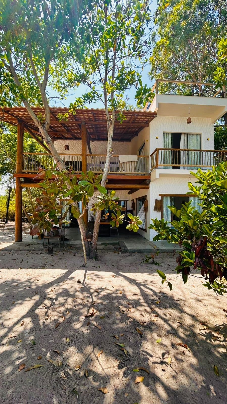 A casa de dois andares, branca, entre as árvores e a areia